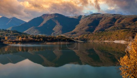 Serene lake reflects golden-hued foliage and evergreens during golden hour, with rolling hills and a dramatic, partly cloudy sky, in an idyllic, remote landscape.