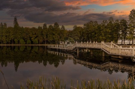 Serene sunrise or sunset over a calm lake with a white, East Asian-inspired bridge, surrounded by lush greenery and reeds, under a vibrant, colorful sky.