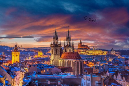 Twilight descends on Prague, casting a golden glow over the Gothic spires of the Church of Our Lady before Tyn and the majestic Prague Castle. Redroofed buildings and flying birds add charm.