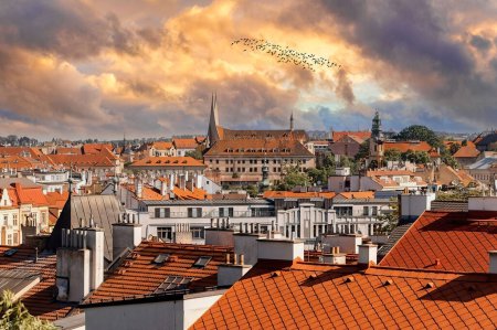 A scenic panorama of Pragues terracotta rooftops, featuring a blend of historical European architecture with church spires and towers rising amidst residential buildings under a partly cloudy sky.