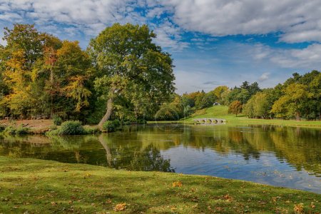 Painshill Park mit dem Serpentinensee und Bogenbrücke und foley mit Eichen und blauem Himmel und weißen Wolken. Painshill Park in Cobham in Surrey England Großbritannien
