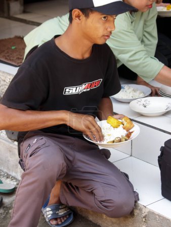 Téléchargez les photos : Un Indonésien local portant un chapeau mange avec sa famille dans un style prêt-à-manger à mains nues - en image libre de droit