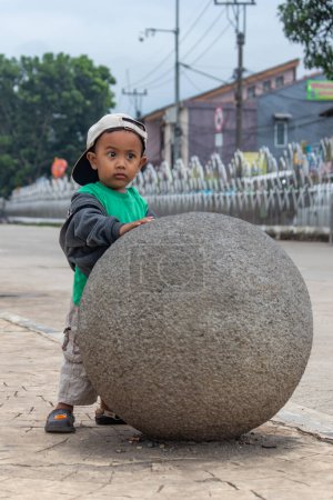Téléchargez les photos : Un jeune enfant, intrigué, explore une grande sphère de pierre, symbolisant l'innocence et la découverte urbaine. - en image libre de droit