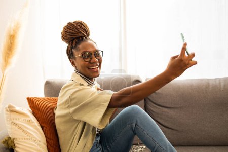 Téléchargez les photos : Souriant jeune afro-américaine téléphone portable à prendre selfie à la maison . - en image libre de droit