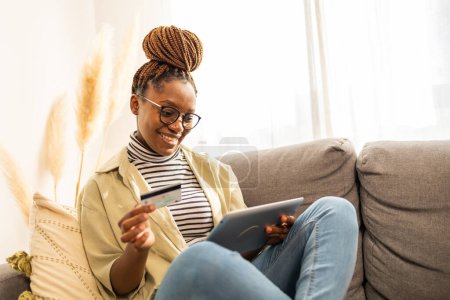 Foto de Joven mujer afroamericana positiva usando tableta digital en casa. Ella está haciendo banca digital, compras en línea. - Imagen libre de derechos