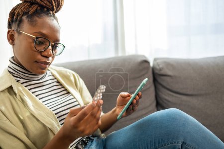 Téléchargez les photos : Souriant jeune femme afro-américaine tablette de téléphone portable à la maison. Elle fait des recherches sur la médecine supplément en ligne - en image libre de droit