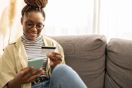 Foto de Sonriente joven mujer afroamericana tableta de teléfono móvil en casa. Ella está haciendo banca digital, compras en línea. - Imagen libre de derechos