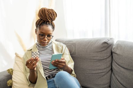 Téléchargez les photos : Souriant jeune femme afro-américaine tablette de téléphone portable à la maison. Elle fait des recherches sur la médecine supplément en ligne - en image libre de droit