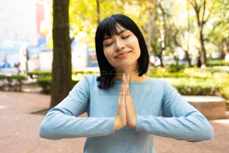 Téléchargez les photos : Portrait de jeune femme asiatique avec les mains en position de prière ou de méditation tout en souriant les yeux fermés à l'extérieur. Photo de haute qualité - en image libre de droit