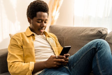 Photo for Portrait of young African American man smiling and using mobile phone at home. High quality photo - Royalty Free Image