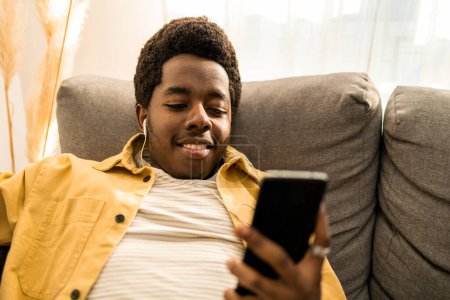 Téléchargez les photos : Portrait de jeune homme afro-américain avec des écouteurs souriant tout en utilisant son téléphone portable. Il est assis confortablement à la maison. Photo de haute qualité. - en image libre de droit