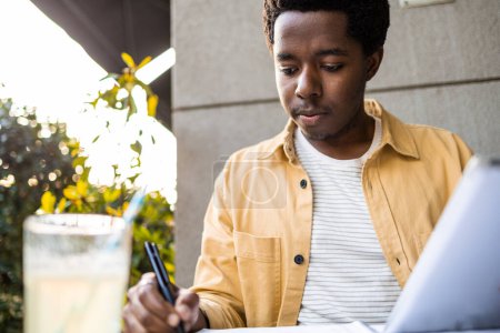Photo for Young African American man having a video call. He is talking from a bar and writing down the while studying online course - Royalty Free Image
