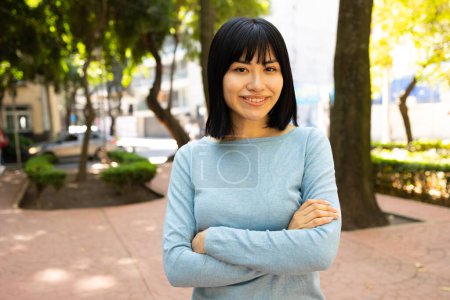 Téléchargez les photos : Portrait de jeune femme asiatique regardant la caméra et souriant à l'extérieur. Elle a croisé les bras. Photo de haute qualité - en image libre de droit