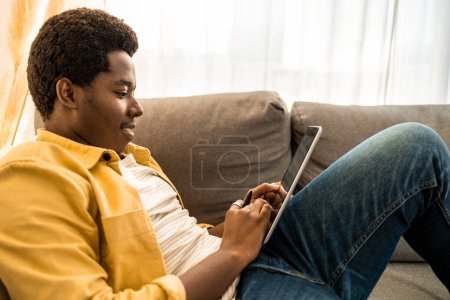 Téléchargez les photos : Vue latérale du jeune homme afro-américain utilisant une tablette numérique à la maison. - en image libre de droit