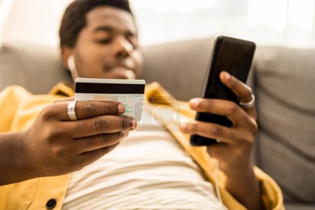 Photo for Portrait of young African American man online paying with credit card in one hand and mobile phone in another. He is sitting at home. High quality photo - Royalty Free Image