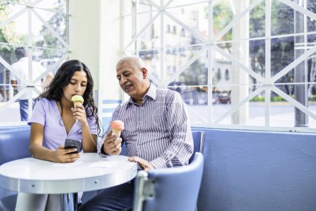 Téléchargez les photos : Adolescente et son grand-père utilisant la technologie tout en appréciant une crème glacée. Photo de haute qualité - en image libre de droit