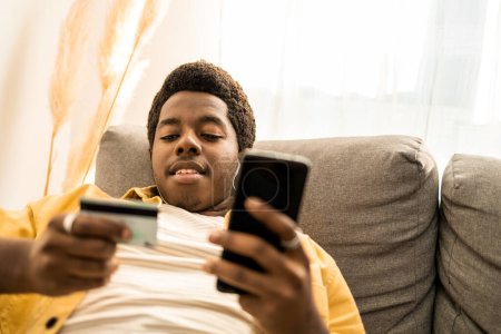 Téléchargez les photos : Portrait de jeune homme afro-américain en ligne payer avec une carte de crédit dans une main et téléphone portable dans une autre. Il est assis à la maison. Photo de haute qualité - en image libre de droit