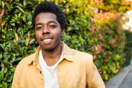 Téléchargez les photos : Portrait de jeune homme afro-américain souriant et regardant la caméra à l'extérieur. Photo de haute qualité - en image libre de droit