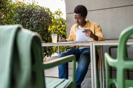 Photo for Young African American man holding tablet and writing down the while studying online course at the bar. - Royalty Free Image