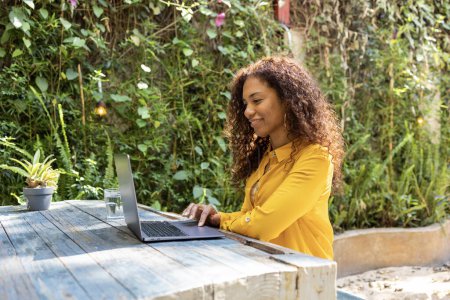 Photo for Side view of Brazilian woman using laptop for freelance work at her backyard. She is wearing yellow sweater. Copy space - Royalty Free Image