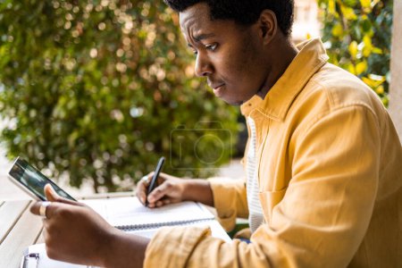 Photo for Side view of young African American man having a video call. He is talking from a bar and writing down the while studying online course - Royalty Free Image