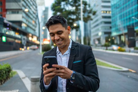 Photo for A portrait of a young Mexican smiling, happy, successful business man, executive walking outside down the street and using mobile phone - Royalty Free Image