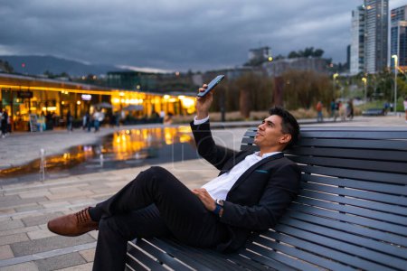Foto de Vista lateral del empresario latino sentado fuera del edificio de oficinas y haciendo una videollamada. Foto al aire libre. Foto de alta calidad - Imagen libre de derechos