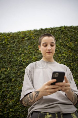 Téléchargez les photos : Portrait de la génération transgenre latine z personne utilisant le téléphone mobile. Il fait du selfie et de la messagerie texte à l'extérieur - en image libre de droit