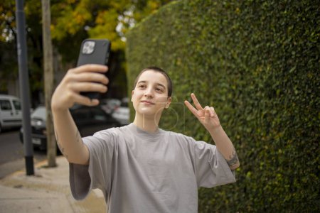 Photo for Portrait of Latin transgender generation z person using mobile phone. He is making selfie and text messaging outdoors - Royalty Free Image