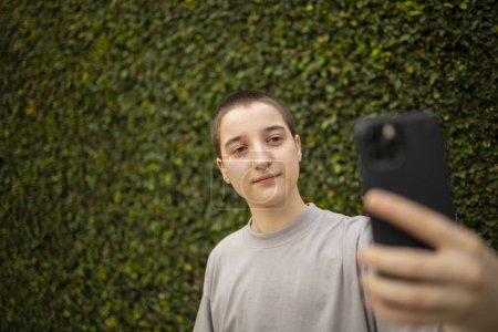Photo for Portrait of Latin transgender generation z person using mobile phone. He is making selfie and text messaging outdoors - Royalty Free Image