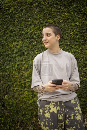 Téléchargez les photos : Portrait de la génération transgenre latine z personne utilisant le téléphone mobile. Il fait du selfie et de la messagerie texte à l'extérieur - en image libre de droit