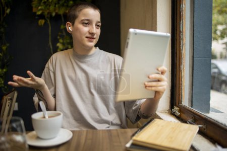 Photo for Latin transgender generation z student using mobile phone, digital tablet at the bar. He is using earphones for video call . High quality photo - Royalty Free Image