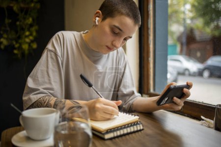 Photo for Latin transgender generation z student using mobile phone, digital tablet at the bar. He is using earphones for video call . High quality photo - Royalty Free Image