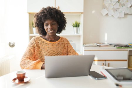 Foto de Sonriente joven afroamericana freelancer utilizando el ordenador portátil en la mesa en la oficina en casa - Imagen libre de derechos