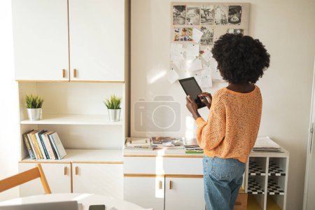 Téléchargez les photos : Vue arrière d'une jeune pigiste noire utilisant une tablette numérique tout en restant debout au bureau à la maison - en image libre de droit
