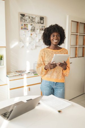 Photo for Happy thoughtful young black woman looking away while holding digital tablet at home office - Royalty Free Image