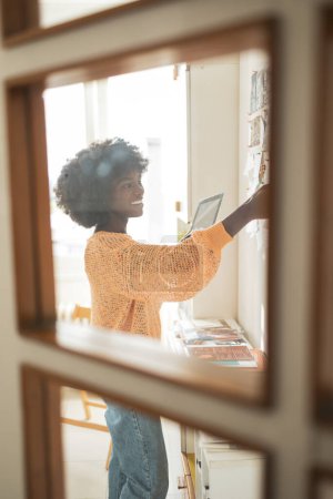 Foto de Feliz joven mujer negra sosteniendo tableta digital mientras trabaja en casa oficina visto a través de la puerta de cristal - Imagen libre de derechos