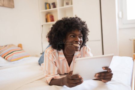 Téléchargez les photos : Pensée jeune femme noire souriant et détournant les yeux tout en tenant tablette numérique dans le lit à la maison - en image libre de droit