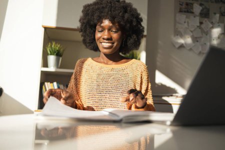 Photo for From below of happy young black woman reading document while working with laptop at table in home office - Royalty Free Image