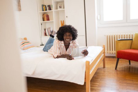 Téléchargez les photos : Pensée heureuse jeune femme afro-américaine regardant loin tout en tenant tablette numérique sur le lit à la maison - en image libre de droit