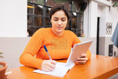 Téléchargez les photos : Jeune femme latine pigiste écriture dans le livre tout en utilisant une tablette numérique au café trottoir - en image libre de droit
