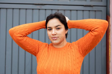 Photo for Portrait of confident young Latin woman in orange sweater with hands behind head standing against gray wall - Royalty Free Image