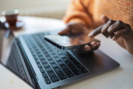 Téléchargez les photos : Femme afro-américaine indépendante méconnaissable utilisant un smartphone avec ordinateur portable à la table dans le bureau à la maison - en image libre de droit