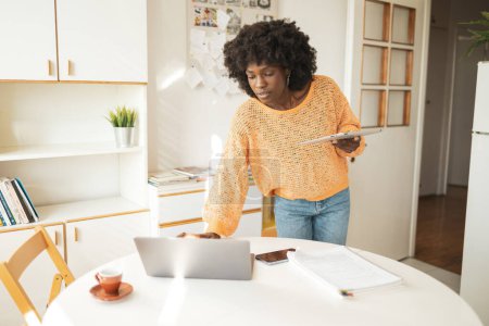 Téléchargez les photos : Jeune pigiste afro-américaine travaillant sur un ordinateur portable tout en tenant une tablette numérique à table au bureau à domicile - en image libre de droit