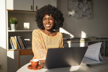 Photo for Thoughtful young black female freelancer looking away while sitting with laptop and documents at table in home office - Royalty Free Image