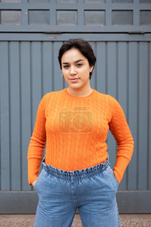 Téléchargez les photos : Portrait de jeune femme latine confiante en pull orange debout avec les mains dans les poches contre le mur gris - en image libre de droit