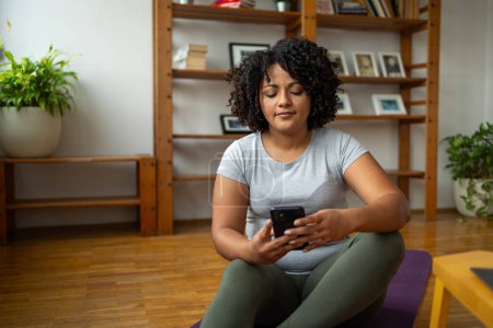 Photo for Young Latin woman in activewear using smartphone while sitting on yoga mat at home - Royalty Free Image
