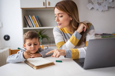 Photo for Confident young blond mother in casual clothes with laptop assisting son with homework at desk at home - Royalty Free Image
