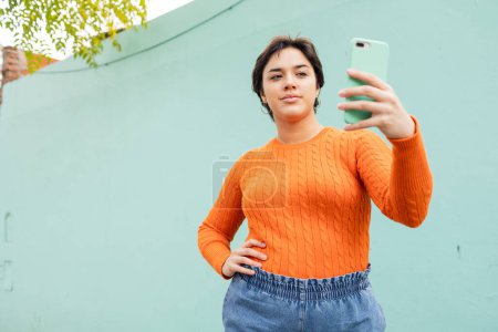 Photo for Confident young Latin woman with hand on hip taking selfie through smartphone while standing against turquoise wall - Royalty Free Image