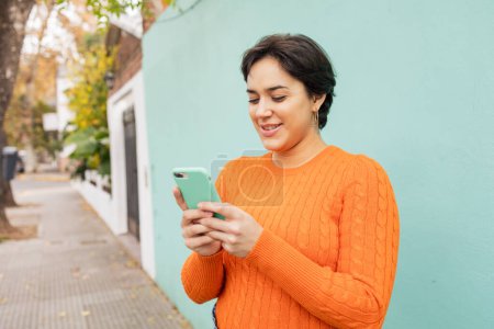 Téléchargez les photos : Heureuse jeune femme latine en pull orange utilisant un smartphone contre un mur turquoise sur le trottoir de la ville - en image libre de droit